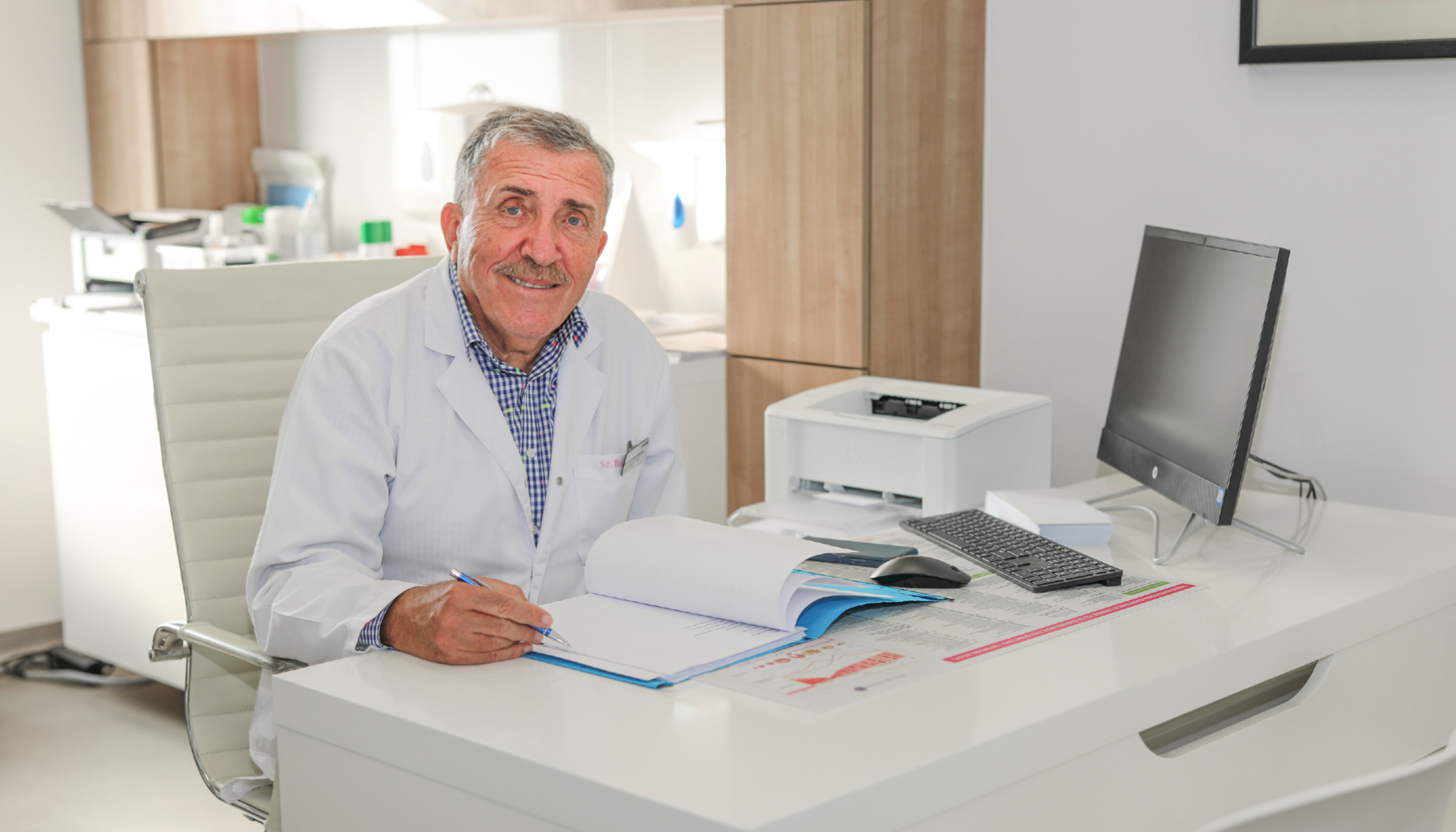 Uśmiechnięty pan siedzący przy biurku przed dokumentami i komputerem, trzymający w dłoni długopis