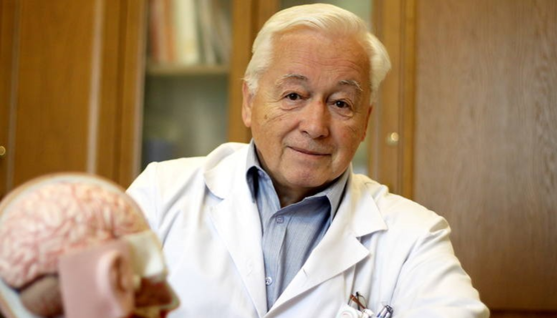 Mężczyzna o siwych włosach i fartuchu lekarskim patrzy w obiektyw aparatu, który zrobił zdjęcie. Przed nim model mózgu