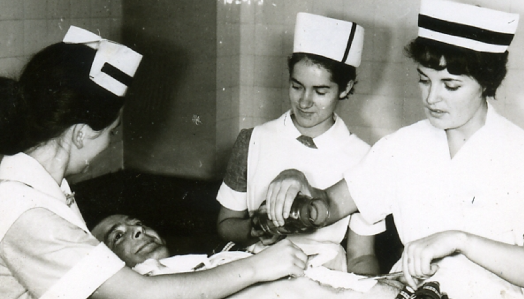 Trzy kobiety w czepkach pielęgniarskich i białych fartuchach w pełnym skupieniu nachylają się nad mężczyzną, prawdopodobnie pacjentem. Jedna trzyma przezroczyste naczynie z nieznaną zawartością.