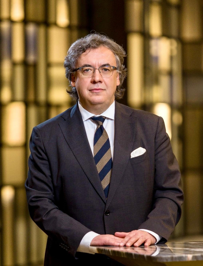 Mężczyzna w garniturze stojący przy stole, w tle rozmyte wnętrze. 