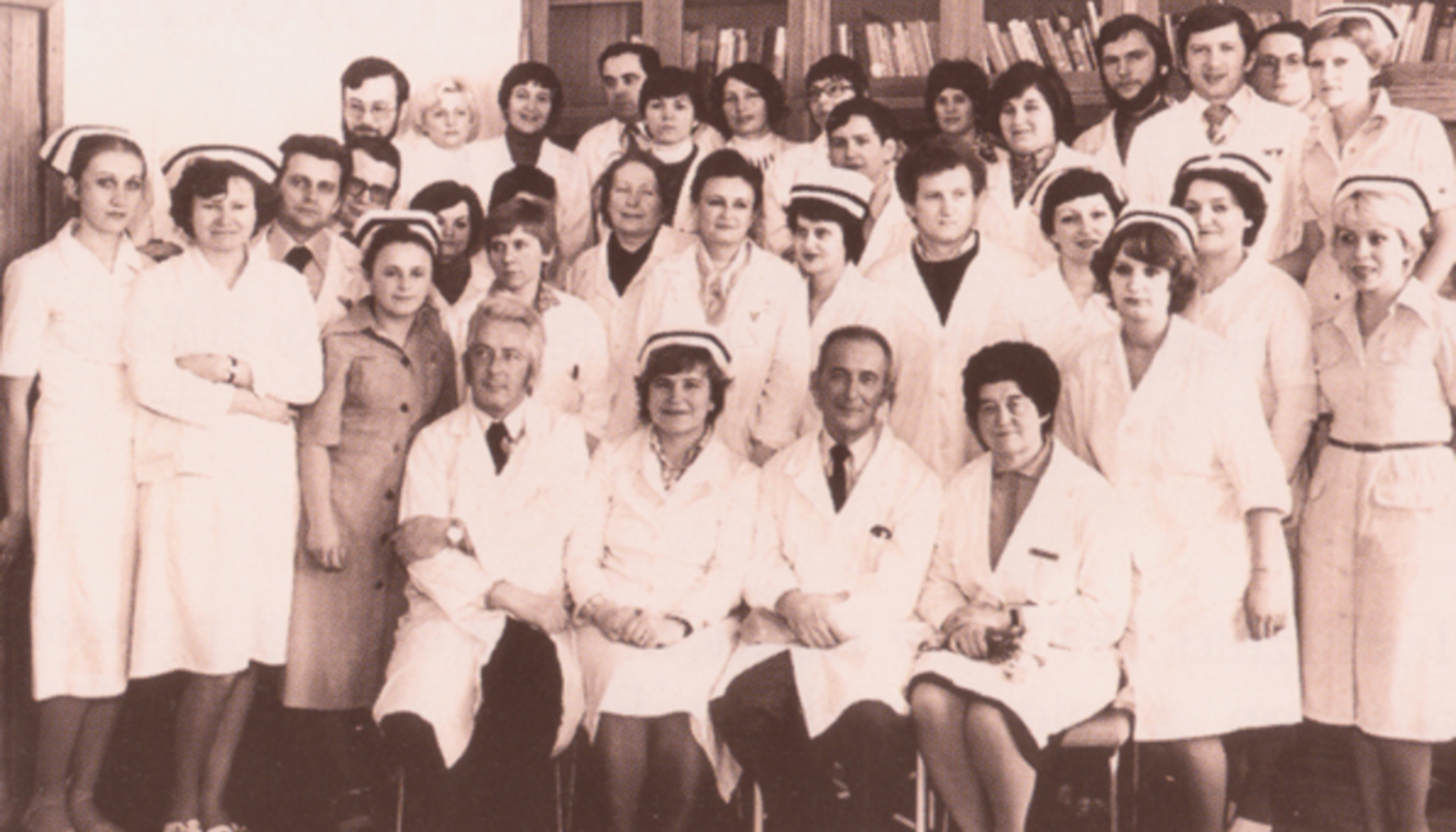 Czarno-białe zdjęcie. Wiele osób w białych fartuchach medycznych, duża część kobiet w czepkach pielęgniarskich, wszyscy ustawieni w rzędach. Z tyłu gablota z książkami.