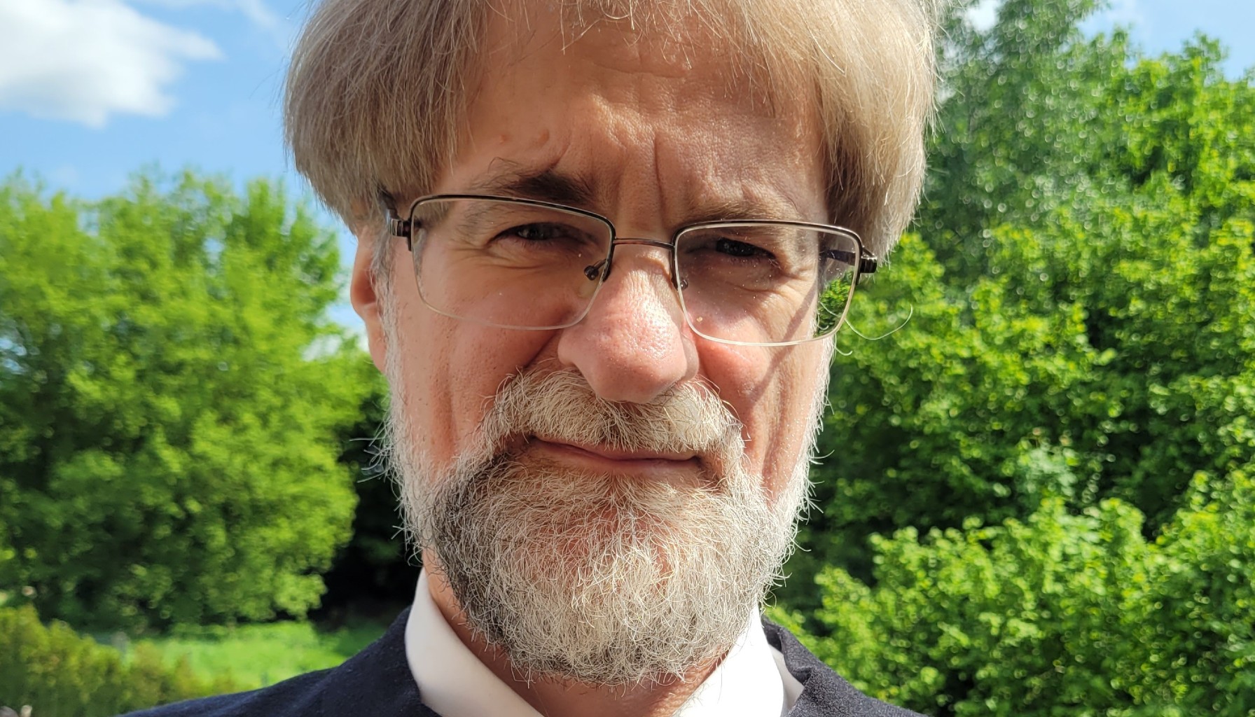 Portret mężczyzny w okularach, z siwą brodą i jasnymi włosami, stojącego na zewnątrz na tle zielonych drzew w słoneczny dzień.