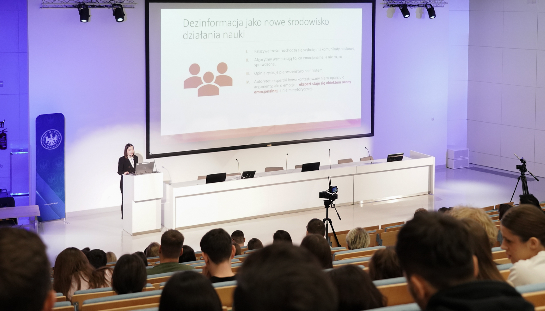 Na zdjęciu widać dużą salę wykładową wypełnioną siedzącymi słuchaczami, patrzącymi w stronę sceny po lewej i na środku fotografii. Na podwyższeniu po lewej stronie stoi kobieta przemawiająca do mikrofonu i pracująca przy laptopie ustawionym na białej mównicy. Obok niej rozciąga się długi biały stół z kilkoma mikrofonami i laptopami, a za nim znajduje się ogromny ekran z wyświetlonym slajdem dotyczącym fałszywych informacji. Po lewej stronie sceny stoi niebieski stojak z logo uczelni, a po prawej stronie ustawiona jest kamera na statywie nagrywająca wydarzenie. Całość oświetlona jest jasnym światłem, a ściany są białe, co nadaje wnętrzu nowoczesny wygląd.