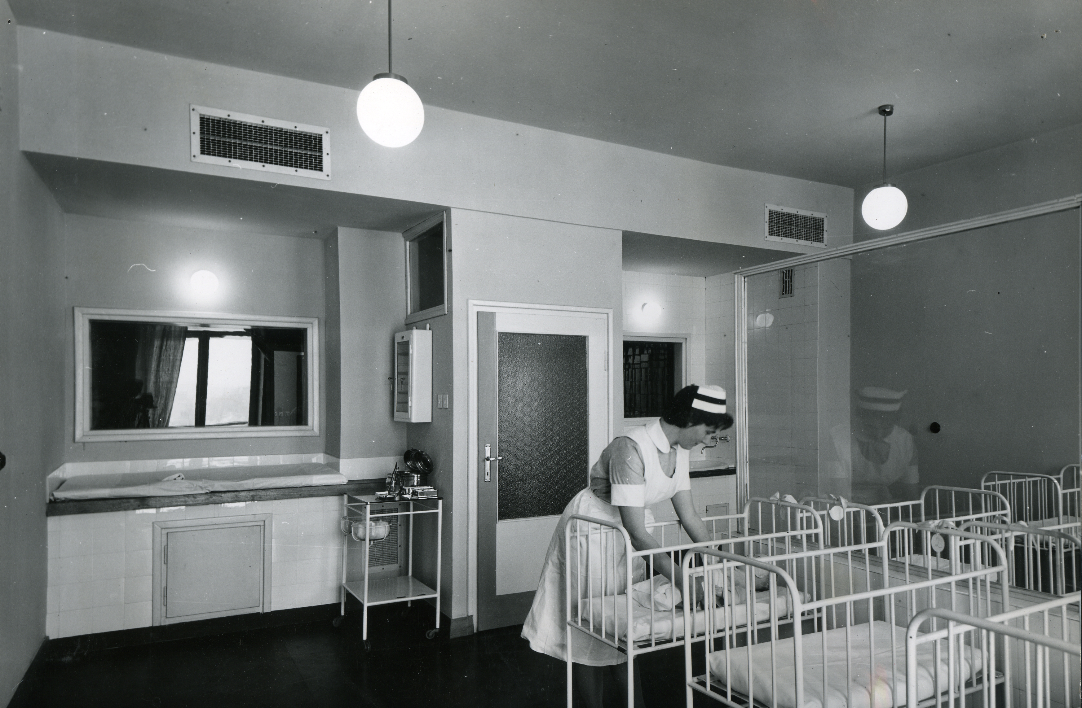 Zdjęcie czarno-białe. Kobieta w czepku schyla się nad łóżeczkami dla noworodków w szpitalnej sali