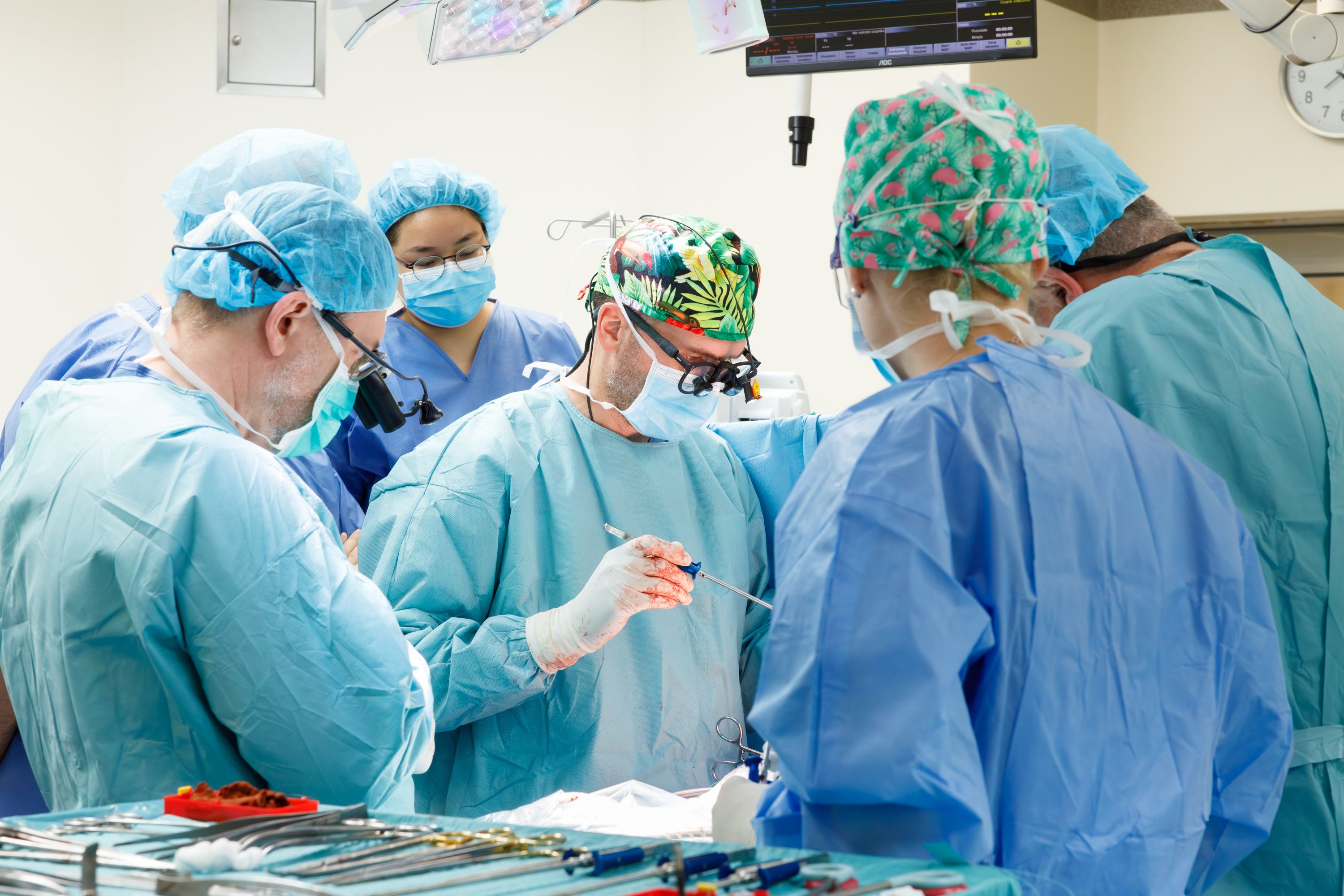 Zdjęcie przedstawia grupę sześciu osób ubranych w jasnoniebieskie lub turkusowe stroje chirurgiczne, pracujących razem wokół stołu operacyjnego w jasnym pomieszczeniu. Wszystkie osoby mają na sobie maski zasłaniające usta i nos oraz czepki chirurgiczne, z czego jeden jest w kolorowy, wzorzysty motyw. Osoby te stoją blisko siebie, skupione na wspólnym punkcie. Centralna postać, lekko pochylona i ubrana w wzorzysty czepek, ma na nosie specjalistyczne okulary lub lupy chirurgiczne i trzyma w dłoni narzędzie. Na pierwszym planie widać stół operacyjny lub stolik z narzędziami pokryty niebieskim materiałem, a na nim leży duża ilość metalowych narzędzi chirurgicznych. W tle, u góry, widoczny jest ekran monitora z cyfrowymi wskaźnikami oraz jasne lampy operacyjne.