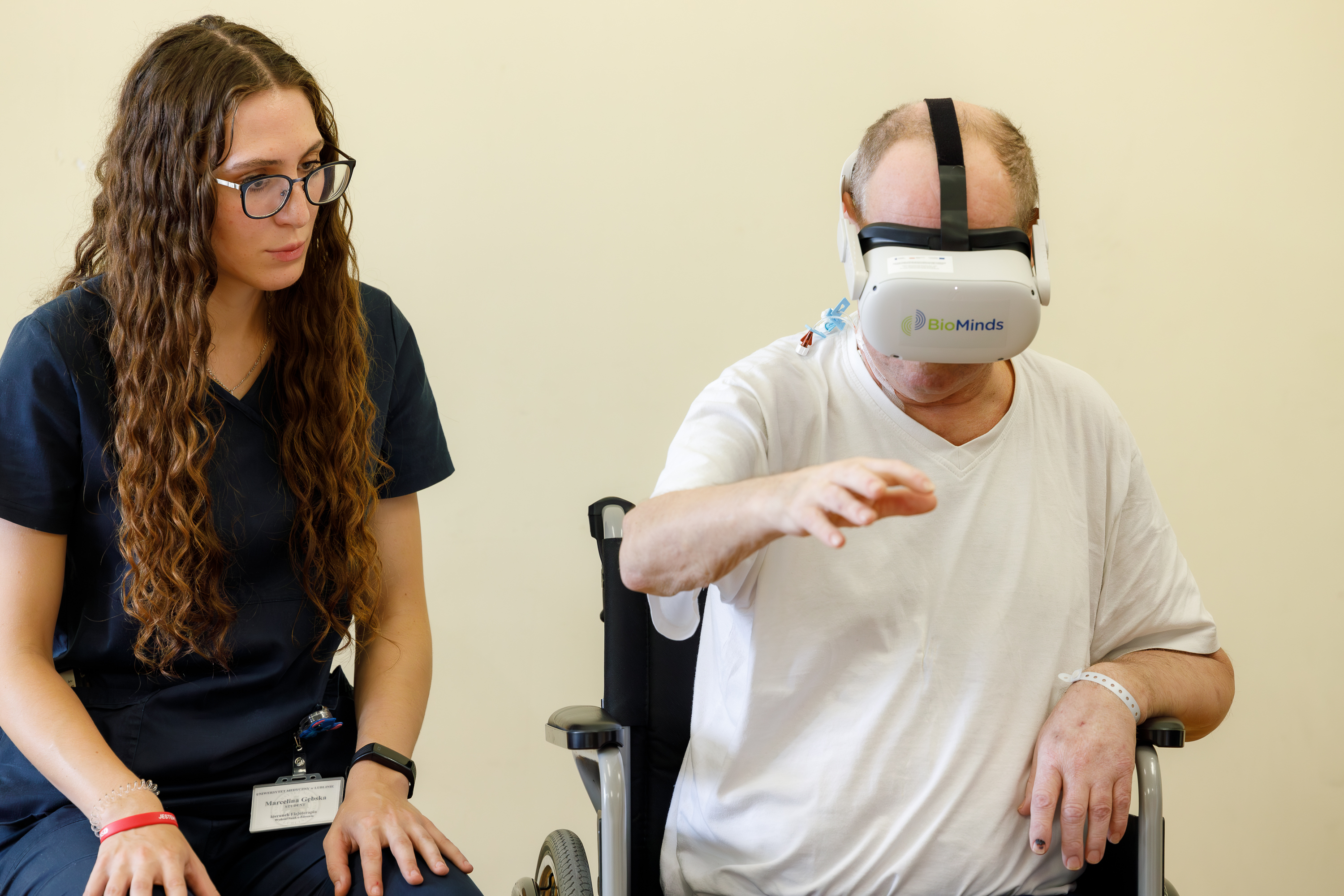 Na zdjęciu widać dwie osoby w jasnym pomieszczeniu na tle beżowej ściany. Po lewej stronie siedzi kobieta z długimi kręconymi brązowymi włosami, ubrana w granatowy strój medyczny i okulary. Ma przy nadgarstku identyfikator i przygląda się drugiej osobie. Po prawej stronie siedzi mężczyzna w białej koszulce, który ma na głowie białe gogle do rzeczywistości wirtualnej z napisem BioMinds. Mężczyzna wyciąga prawą rękę przed siebie, wykonując jakiś gest związany z ćwiczeniami w wirtualnej rzeczywistości. Siedzi na czarnym krześle z podłokietnikami. Kobieta obserwuje go uważnie, co sugeruje, że prowadzi sesję terapeutyczną z wykorzystaniem technologii gogli VR.