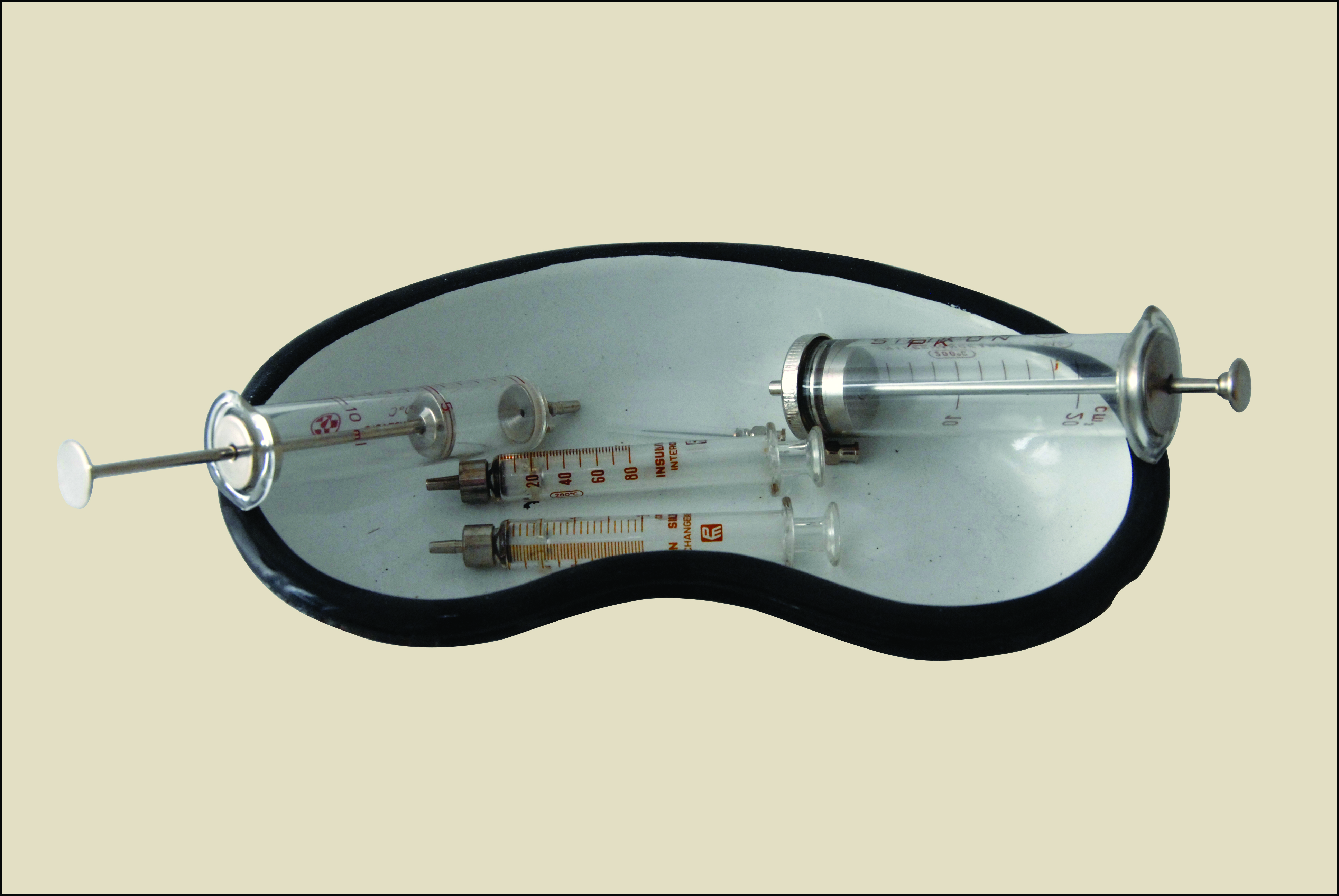 Na zdjęciu widać naczynie w kształcie nerki, białe z czarnymi konturami, w środku znajdują się różnie wyglądające strzykawki starego typu, szklane, z zaznaczoną skalą i metalowymi elementami.