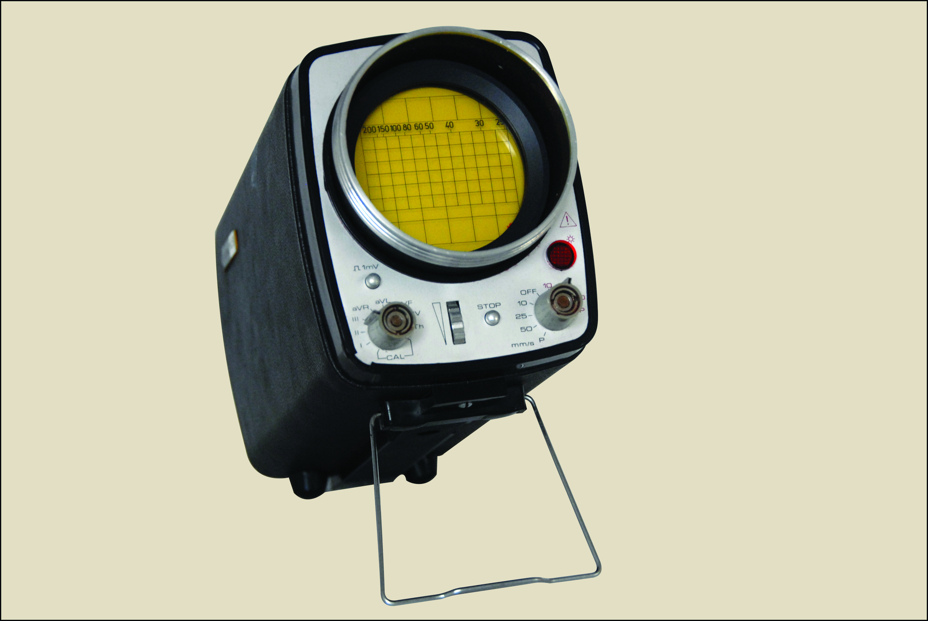 Zdjęcie przedstawia przenośne urządzenie elektroniczne w czarnej prostokątnej obudowie stojące na cienkiej metalowej podpórce z drutu na jednolitym beżowym tle. Urządzenie jest lekko pochylone do tyłu co eksponuje jego srebrny panel przedni. W górnej części panelu dominuje duży okrągły ekran otoczony wystającą czarną osłoną. Wyświetlacz ma żółte tło z naniesioną czarną siatką pionowych i poziomych linii oraz rzędem cyfr przy górnej krawędzi. Poniżej ekranu znajduje się panel sterowania na którym po prawej stronie umieszczono okrągłą czerwoną lampkę z kratką. W dolnej części panelu wystają dwa symetrycznie rozmieszczone metalowe gniazda jedno po lewej i jedno po prawej stronie. Pomiędzy nimi znajduje się wąskie pionowe pokrętło oraz mały srebrny przycisk. Na powierzchni panelu widoczne są również drobne czarne napisy i symbole.