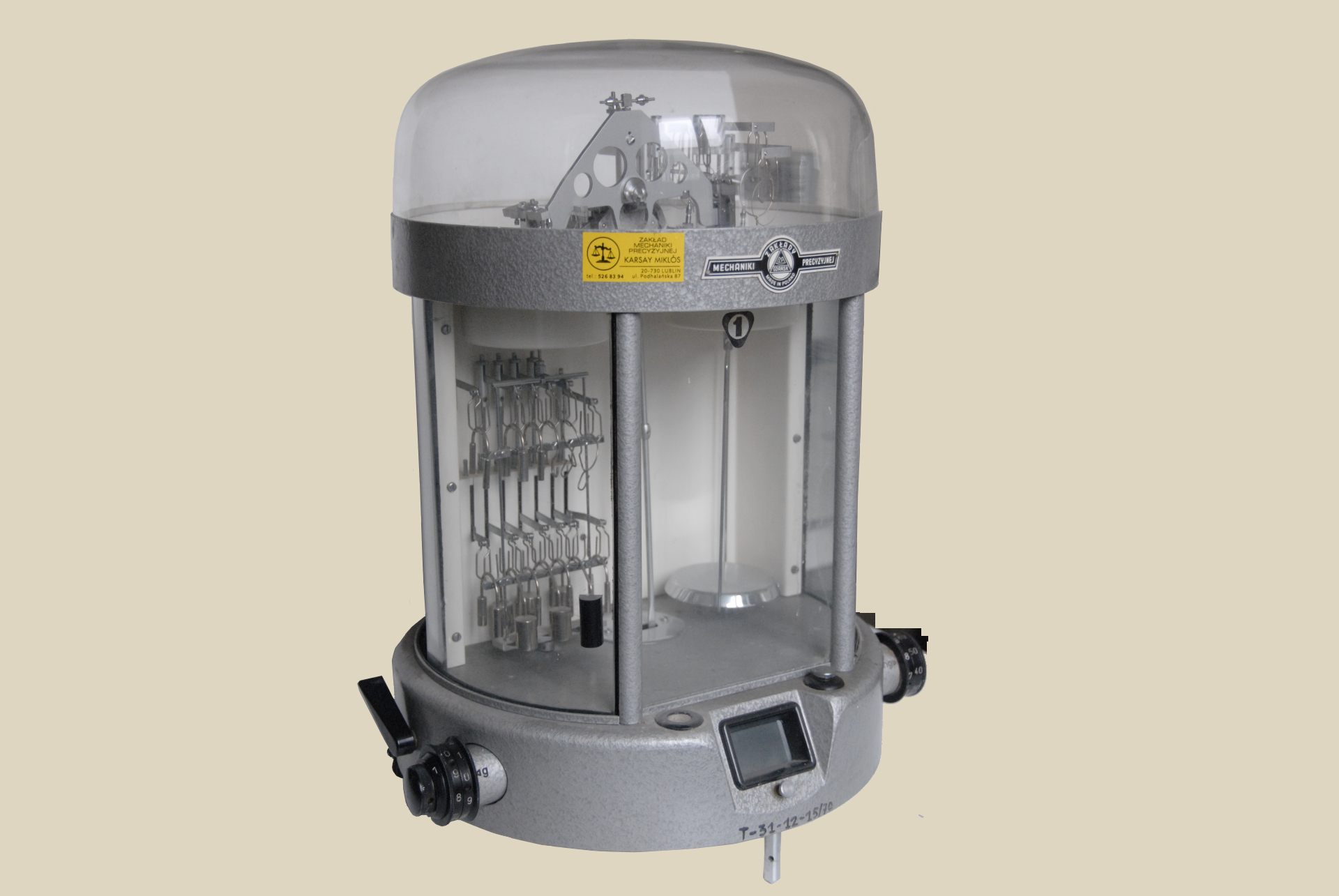 Na zdjęciu widać urządzenie medyczne w kolorze szarym o cylindrycznym kształcie, umieszczone na jasnym tle. Sprzęt ma przezroczystą kopułę na górze, przez którą widać wewnętrzne metalowe mechanizmy i rurki. Część środkowa urządzenia jest otoczona szarą opaską z naklejonymi żółtymi i białymi etykietami z napisami. Poniżej znajduje się przezroczysta komora, w której po lewej stronie widoczne są pionowo ułożone metalowe elementy przypominające wieszaki lub uchwyty, a po prawej znajduje się pusta przestrzeń z pionowym elementem. Na dolnej części urządzenia znajdują się czarne pokrętła po obu stronach oraz mały prostokątny wyświetlacz lub panel kontrolny z przodu. U dołu widać numer identyfikacyjny urządzenia. 