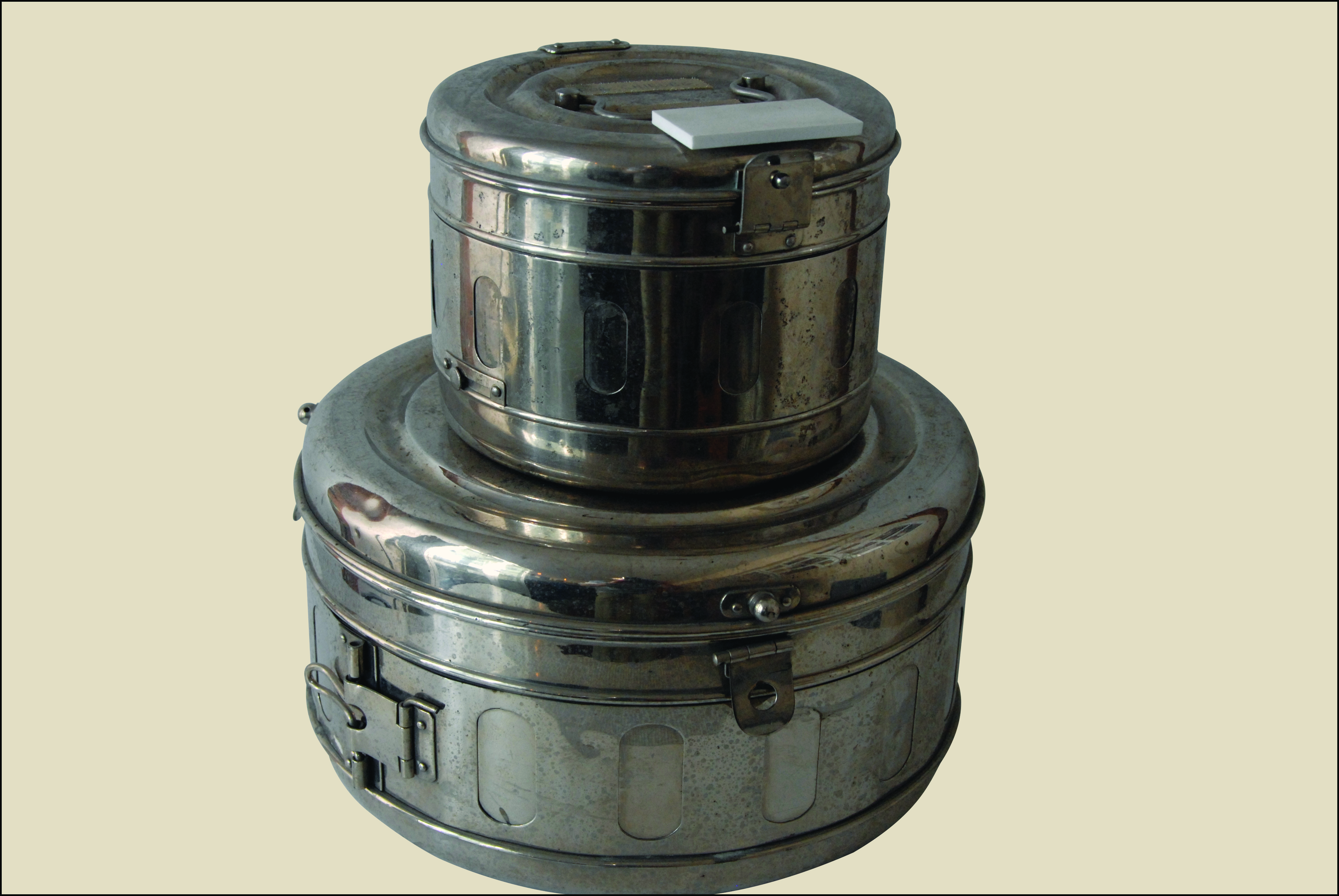 Na zdjęciu widać dwie metalowe puszki z zatrzaskami. Jedna puszka jest większa, o szerokiej osi, druga mniejsza. Na górze mniejszej puszki leży prostokątny kartonik.