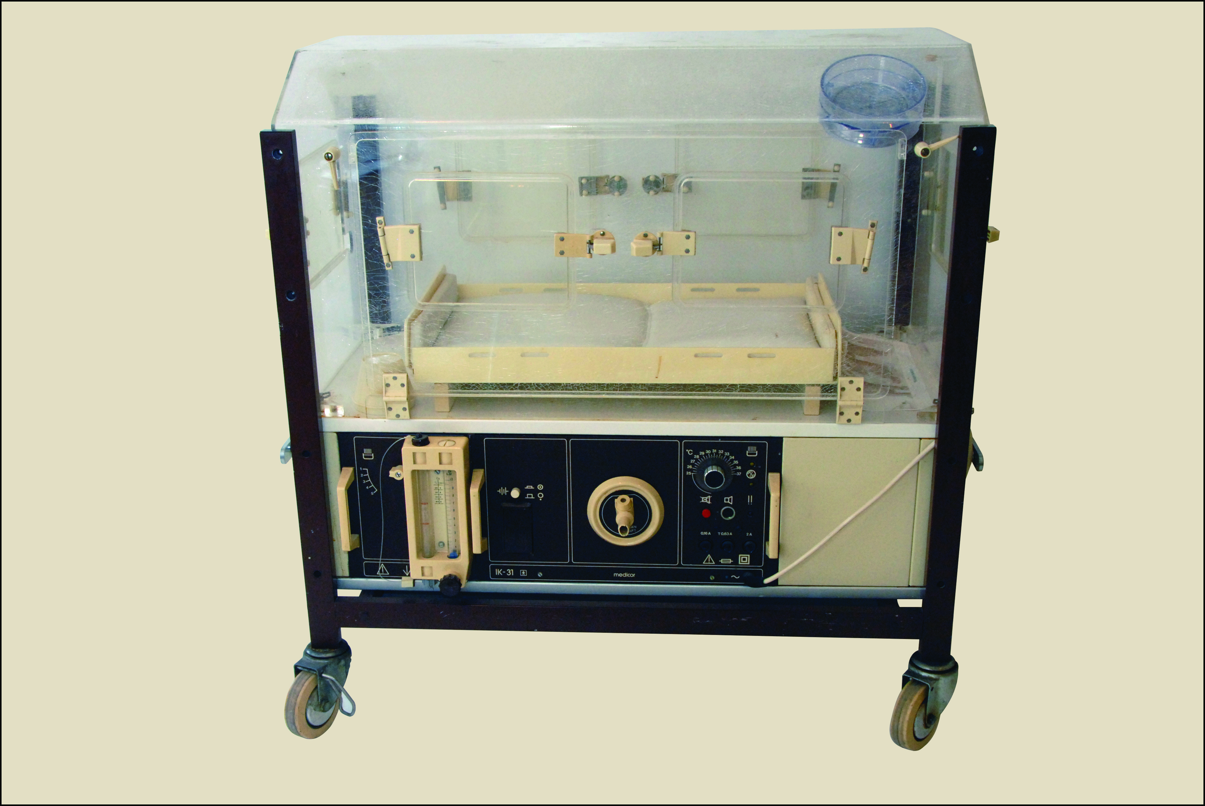 Na zdjęciu widać dużą, prostopadłościenną maszynę na kółkach, przypominającą szafkę, ale z przezroczystą, górną częścią. Całość jest osadzona na metalowym stelażu, pomalowanym na ciemny kolor.  W górnej części znajduje się duża, przezroczysta obudowa, wyglądająca jak prostopadłościenne pudełko z przezroczystego plastiku lub szkła. W środku jest widoczne małe, jasne łóżeczko, które wygląda jak stelaż z prostokątnym materacem. Jest ono podparte na drewnianych lub plastikowych elementach. Na górze obudowy, po prawej stronie przymocowana jest przezroczysta miseczka. Dolna część maszyny jest nieprzezroczysta, ma kolor beżowy i czarny. Widać na niej różne, okrągłe pokrętła, przełączniki, tarcze z cyframi oraz małe lampki kontrolne. Na tej płycie znajduje się też mały, prostokątny ekran, a obok niego, po lewej stronie, widoczna jest pionowa, przezroczysta rurka ze skalą.