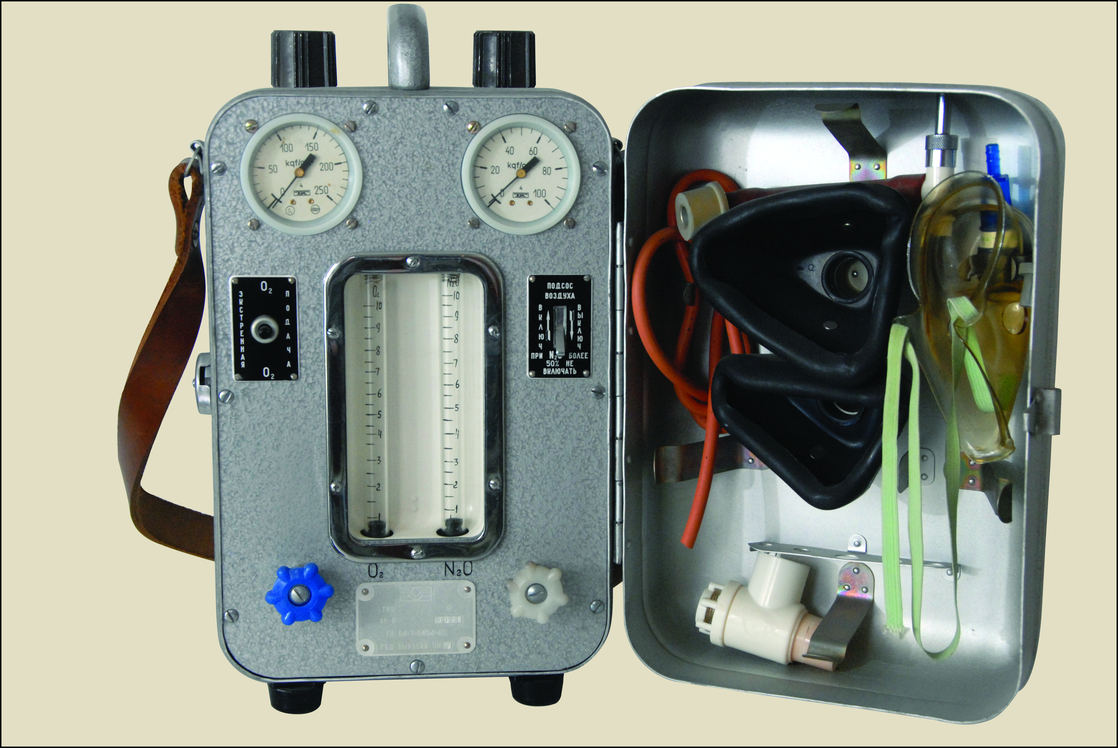 Na zdjęciu widoczna jest metalowa, przenośna skrzynka, przypominająca walizkę narzędziową lub apteczkę polową, otwarta na prawą stronę. Główna część skrzynki, w kolorze szarym, zawiera zestaw wskaźników i elementów kontrolnych. Na górze umieszczono dwa okrągłe zegary z białymi tarczami i czarnymi cyframi, a pośrodku duży, pionowy, przezroczysty element ze skalą. Poniżej znajdują się niebieskie i białe pokrętła oraz różne przełączniki. W górnej części walizki znajduje się metalowy uchwyt do przenoszenia, a z lewej strony wystaje brązowy pasek na ramię. Wnętrze otwartej pokrywy, po prawej stronie, służy jako schowek i wypełniają je elastyczne elementy: duża, czarna, gumowa forma przypominająca maskę na twarz, zwinięte pomarańczowe i przezroczyste rurki oraz kilka mniejszych, białych i przezroczystych plastikowych części.