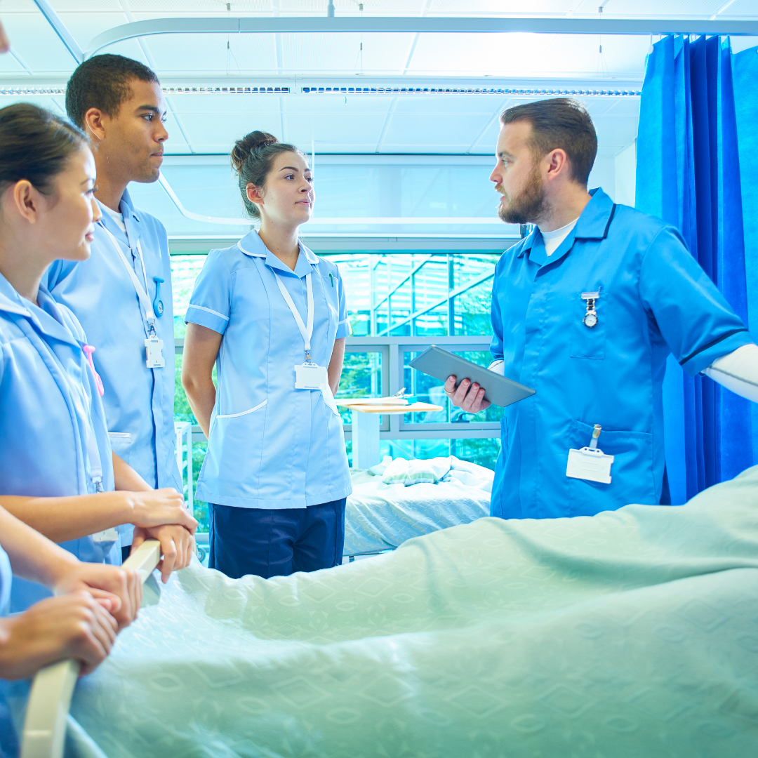 Grupa pielęgniarzy i pielęgniarek w niebieskich uniformach uczestniczy w szkoleniu przy łóżku pacjenta w szpitalnej sali. Jeden z nich, trzymając tablet, prowadzi rozmowę edukacyjną z pozostałymi osobami.