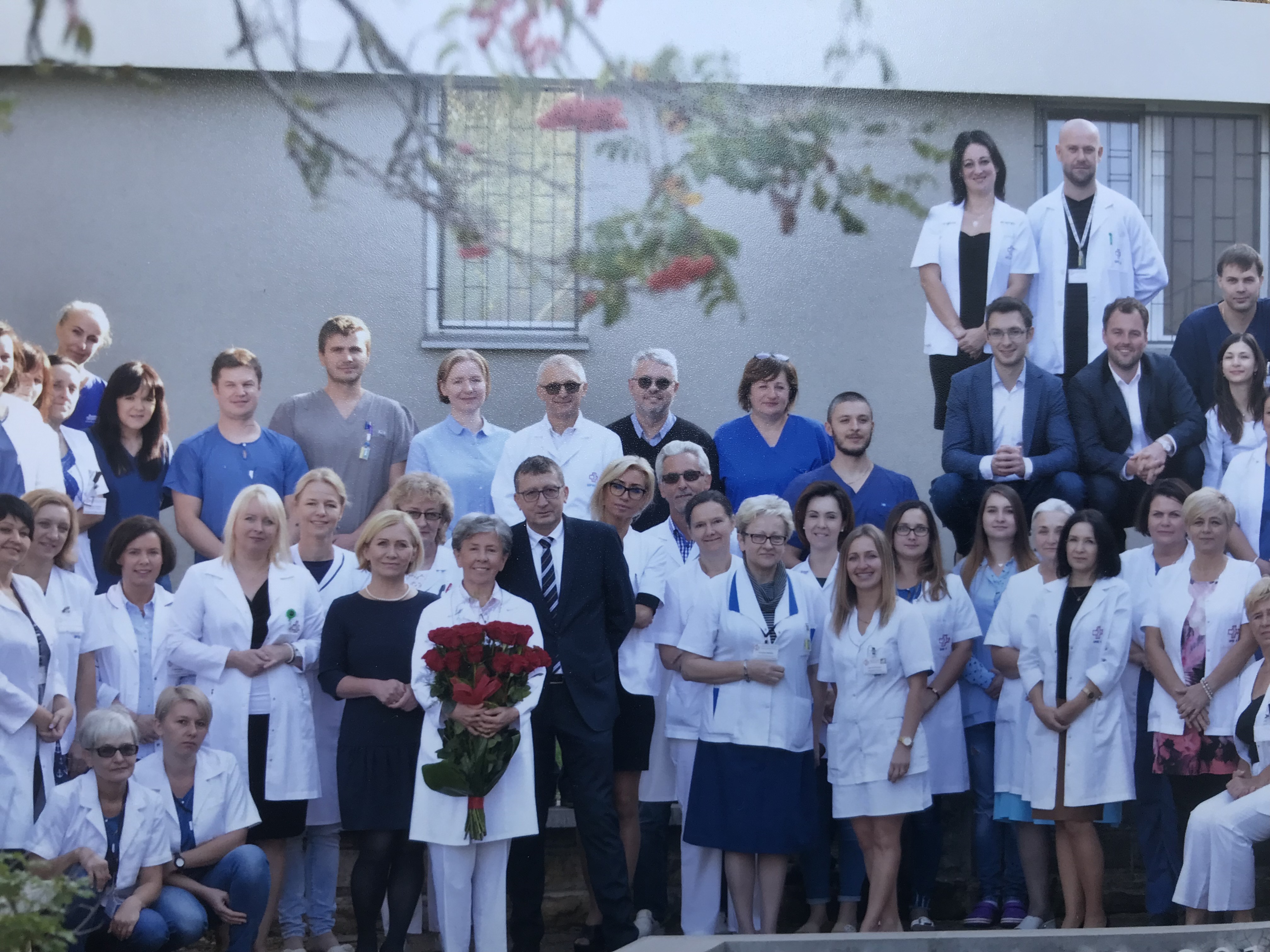 Duża grupa os&oacute;b w fartuchach lekarskich lub garniturach pozująca na tle ściany budynku, z przodu widać niewyraźne gałęzie drzewa.