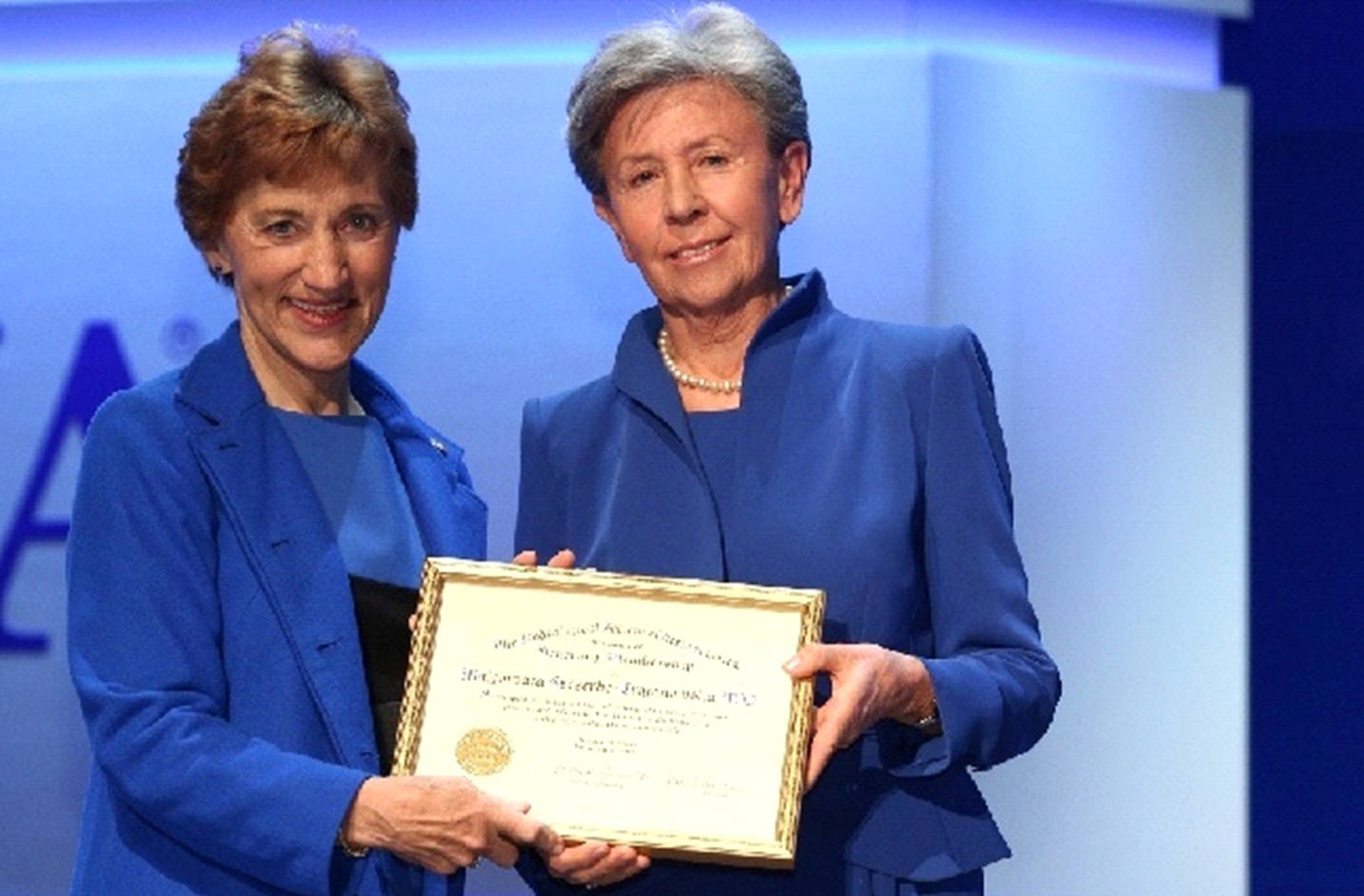 Dwie eleganckie, uśmiechnięte kobiety na granatowym tle, trzymające dyplom o nieczytelnej treści w złotej ramie.