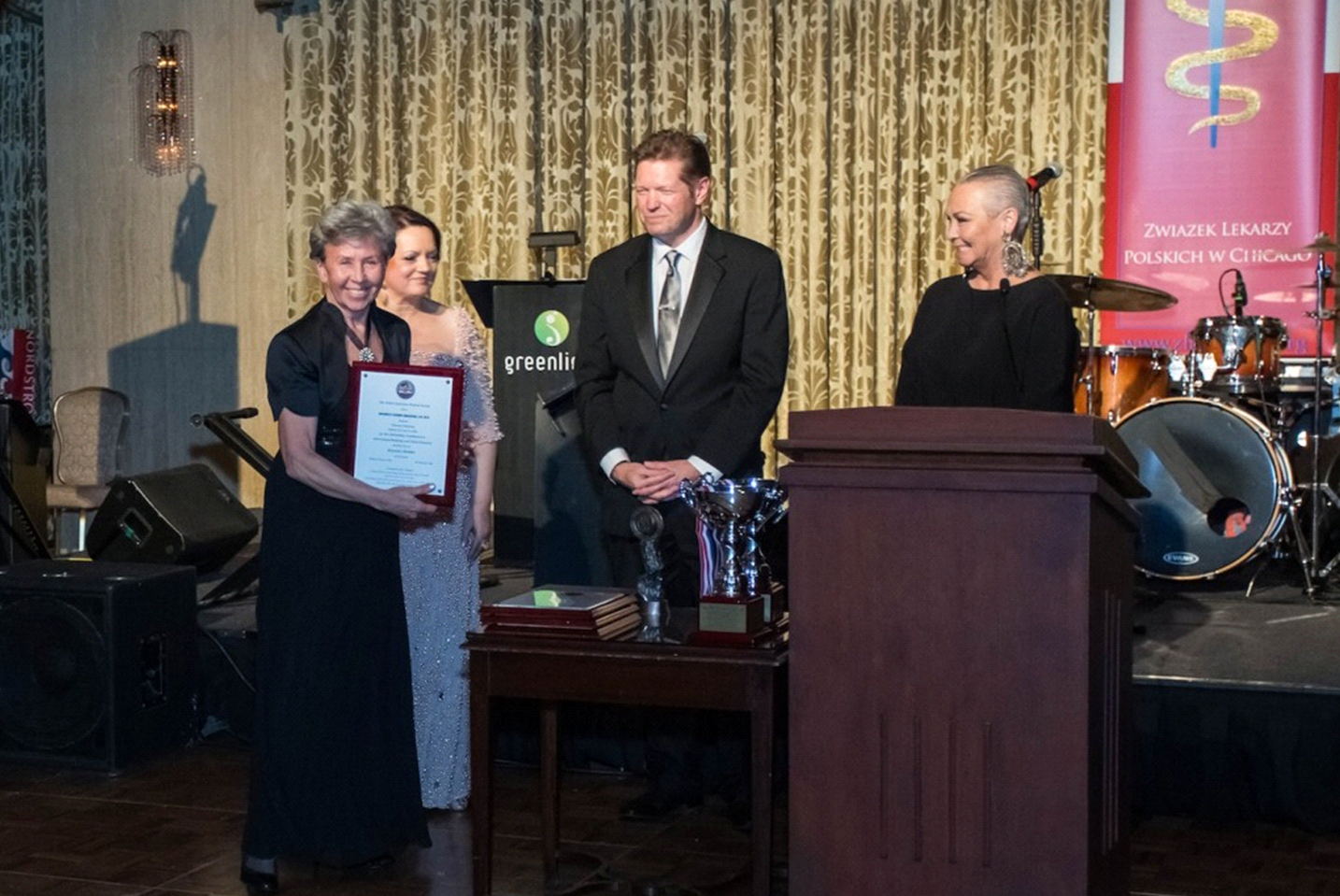 Na zdjęciu znajduje się uroczysta scena wręczenia jakiegoś wyr&oacute;żnienia. Na pierwszym planie, po lewej stronie, starsza kobieta z kr&oacute;tkimi siwymi włosami uśmiecha się i trzyma w rękach dużą, oprawioną w ramkę dyplomową tablicę &mdash; wygląda na to, że właśnie otrzymała wyr&oacute;żnienie. Ma na sobie czarną, elegancką suknię. Za nią stoi kobieta w jasnej, błyszczącej sukni, kt&oacute;ra także się uśmiecha. Obok nich, na środku zdjęcia, stoi mężczyzna w czarnym garniturze i krawacie, a po prawej &mdash; kobieta z ogoloną głową, w czarnej sukni, stojąca przy m&oacute;wnicy. W tle widoczny jest zestaw perkusyjny oraz czerwony baner z napisem &bdquo;Związek Lekarzy Polskich w Chicago&rdquo;. Panuje podniosła atmosfera, a scena jest dobrze oświetlona.