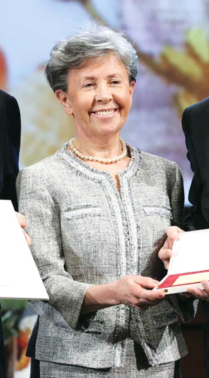 Kobieta uśmiechnięta, elegancko ubrana w komplet z żakietem, z naszyjnikiem z pereł na szyi trzyma otwarte płaskie pudełko z trudną do rozpoznania zawartością.