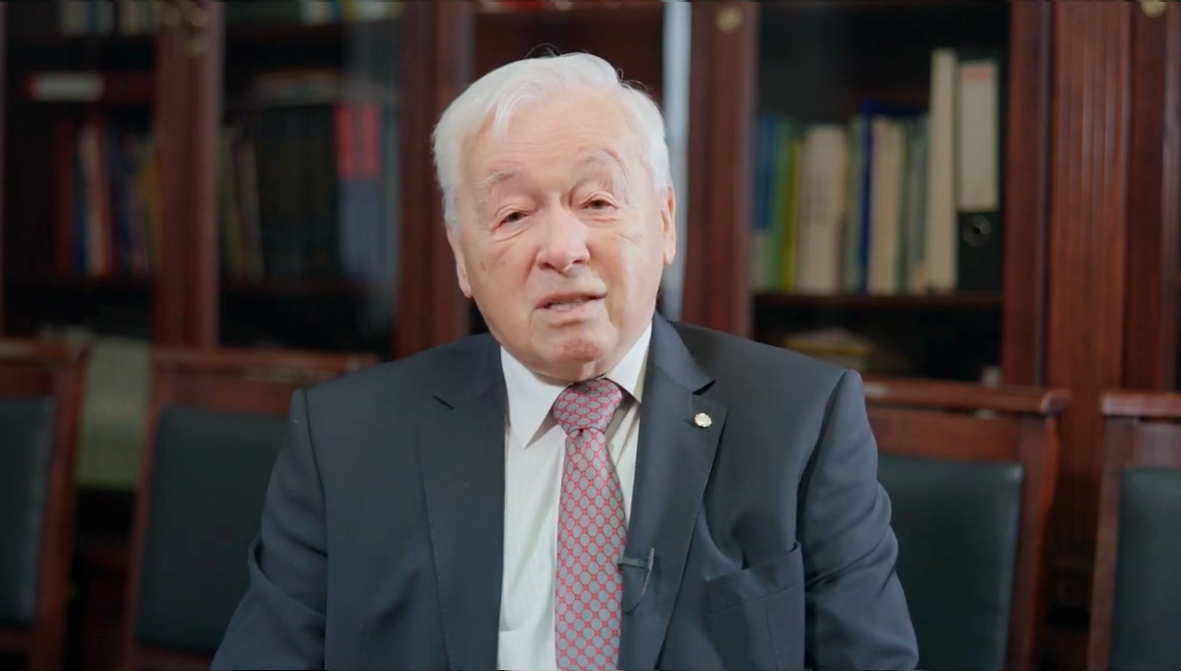 Siwy mężczyzna w garniturze widziany od twarzy do klatki piersiowej, na tle p&oacute;łek z książkami.