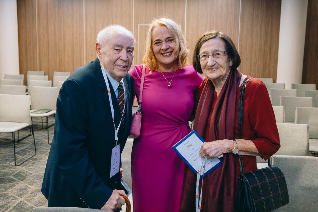 Trzy uśmiechnięte osoby w sali z krzesłami. W środku kobieta w r&oacute;żowej sukni obejmuje po swojej prawej stronie starszego mężczyznę, a po swojej stronie starszą kobietę. Wszyscy są uśmiechnięci.