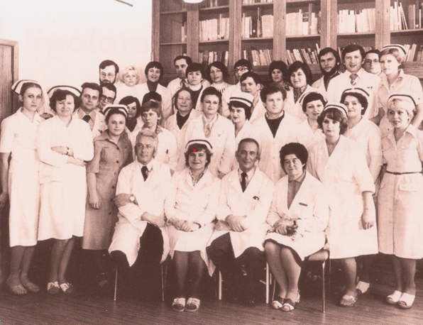 Czarno-białe zdjęcie. Wiele os&oacute;b w białych fartuchach medycznych, duża część kobiet w czepkach pielęgniarskich, wszyscy ustawieni w rzędach. Z tyłu gablota z książkami.